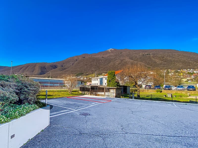 Empty parking lot with multiple parking spots, surrounded by green grass and bushes, mountains in the background.