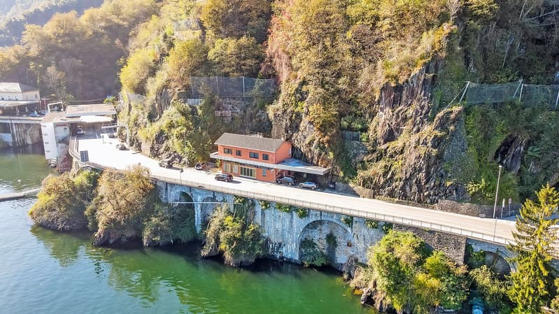 Bridge house with bridge, orange house, cars parked, trees, greenery, rocks, river