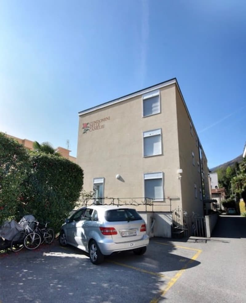 Apartment building, beige walls, windows with white shutters, car parked in front, bicycles leaning against a bush