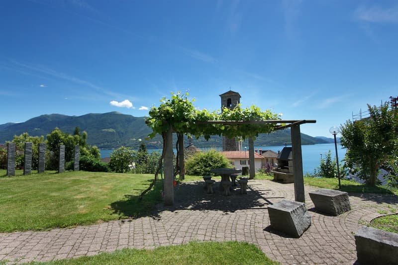 Garden with a table and chairs under a pergola, brick floor, grassy lawn, mountains, lake, and bell tower in the background