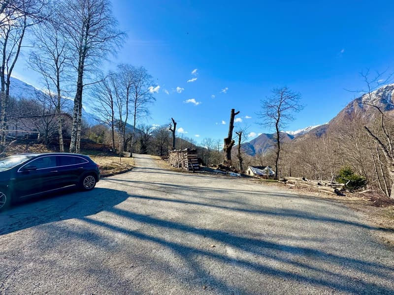 Country road with a car parked, surrounded by bare trees, mountains, and blue sky with some clouds.