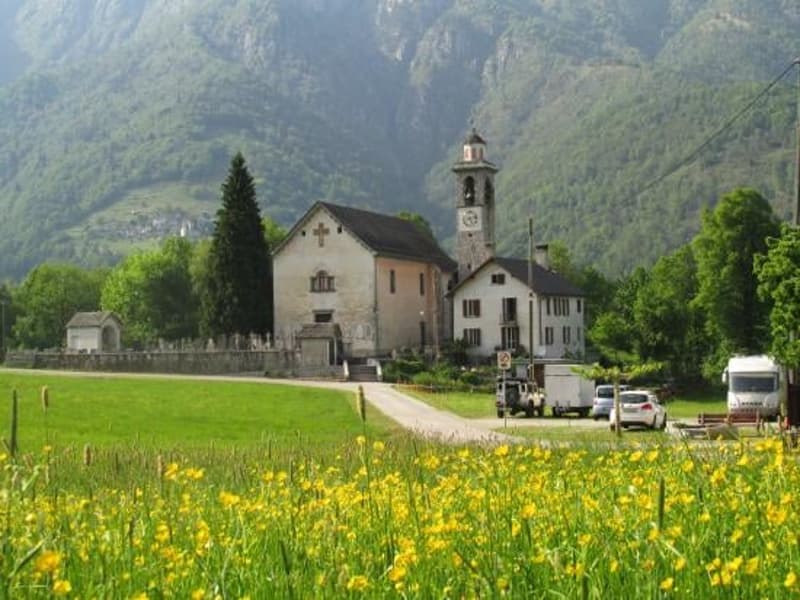 The image shows a picturesque village nestled in a mountainous landscape. The village features a church with a bell tower, surrounded by traditional buildings and a grassy field filled with yellow flowers. There are also some parked vehicles visible in th