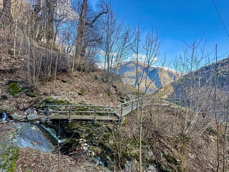 The image shows a scenic outdoor landscape with a small wooden bridge crossing over a stream or creek. The surrounding area is covered in bare trees and vegetation, with a blue sky visible in the background. The landscape appears to be in a mountainous or