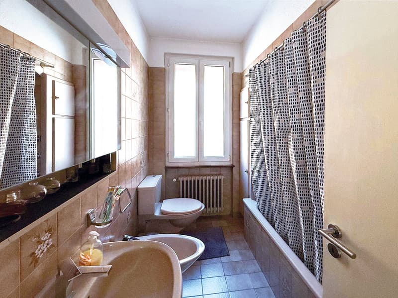 Tiled bathroom with a bathtub, toilet, vanity, and patterned curtains. There is a washing machine visible in the room.