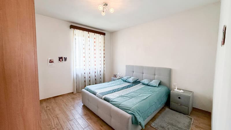Spacious bedroom with hardwood flooring, white walls, a large bed with a gray upholstered headboard, bedside tables, and curtained windows.