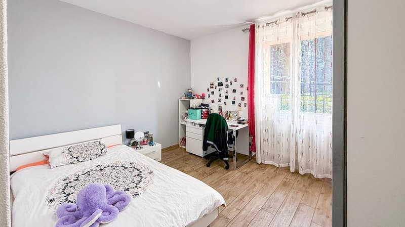 Spacious bedroom with large windows, hardwood flooring, white walls, a bed with patterned bedding, a desk with shelves, and a purple stuffed animal on the bed