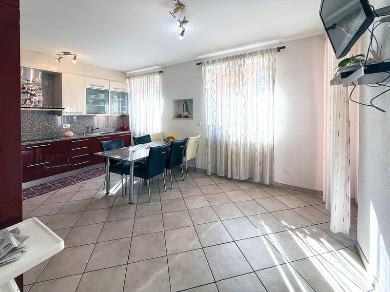 Open-concept kitchen and dining area with tile flooring, burgundy cabinets, granite countertops, and a dining table with chairs. The room has large windows and curtains, and a dishwasher is visible in the kitchen area.