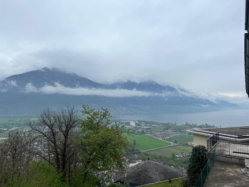 The image shows a scenic view of a mountainous landscape with clouds covering the peaks. There are trees in the foreground and a town or city visible in the valley below. The overall scene appears to be a picturesque view from a high vantage point, with t