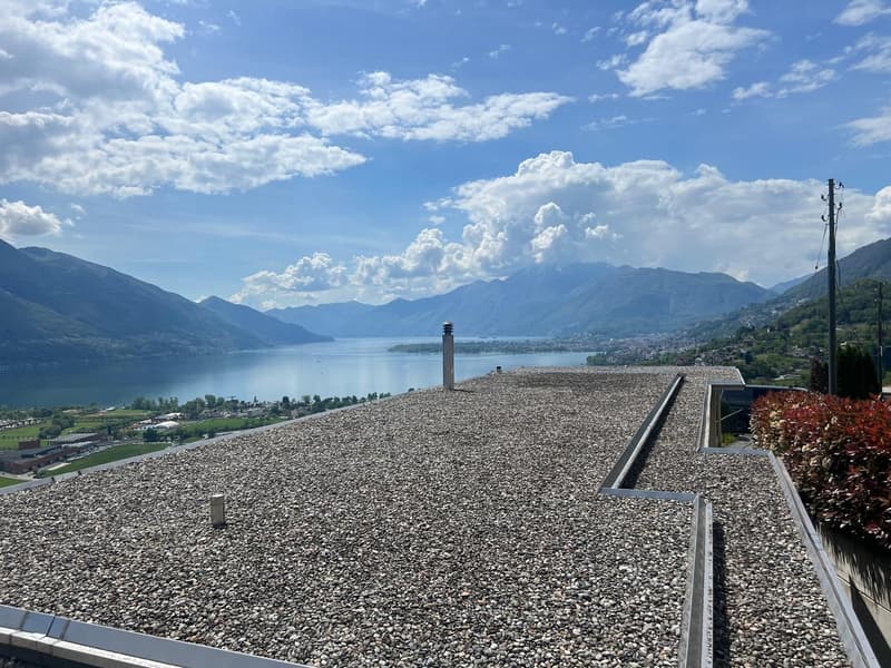The image shows a scenic view of a lake surrounded by mountains. The foreground features a large gravel or pebble-covered roof or terrace, with a chimney or tower structure in the center. The lake and mountains in the background are visible through the cl