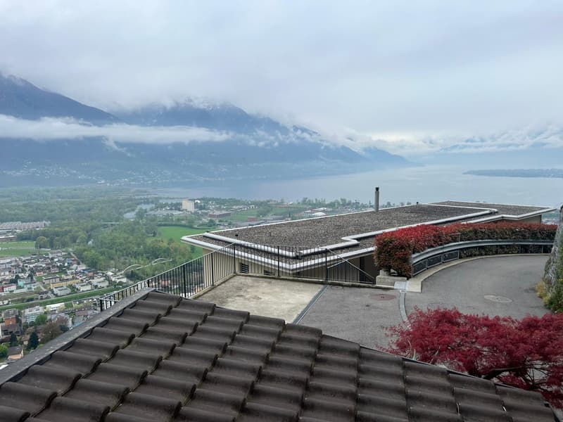 The image shows a scenic view of a mountainous landscape with a town or city in the valley below. The foreground features a building with a slanted roof and a balcony or terrace overlooking the view. The building appears to be situated on a hillside, prov