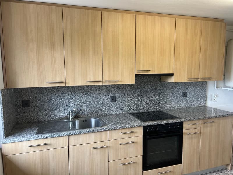 The image shows a modern kitchen with light wood cabinets, a granite countertop, a stainless steel sink, and a built-in oven. The kitchen has a clean and minimalist design.