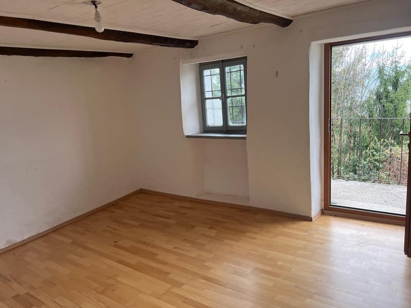 This image shows a room with hardwood floors, white walls, and exposed wooden beams on the ceiling. The room has a large window that provides a view of the outdoor greenery. The space appears to be empty, ready for furnishing and decorating.