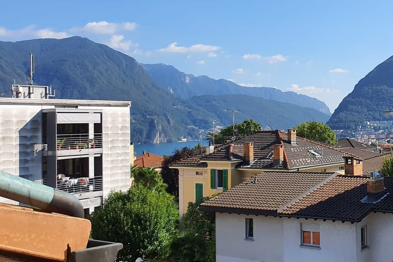 The image shows a scenic view of a town or city nestled between mountains. The buildings have red tile roofs and there are balconies visible on some of the structures. The landscape is lush with greenery, and the lake or body of water in the background ad
