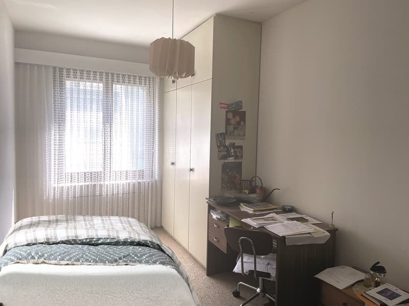 The image shows a bedroom with a bed, a wardrobe, and a desk with various items on it. The room has large windows with sheer curtains, providing natural light. The overall decor appears simple and minimalist.