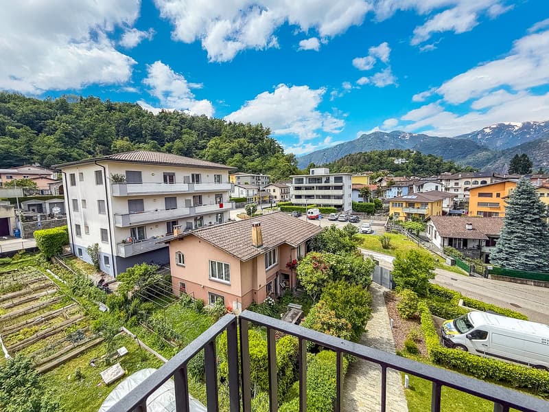 The image shows a residential area with multi-story apartment buildings, surrounded by lush greenery and mountains in the background. The buildings have balconies and the area appears well-maintained with gardens and landscaping.