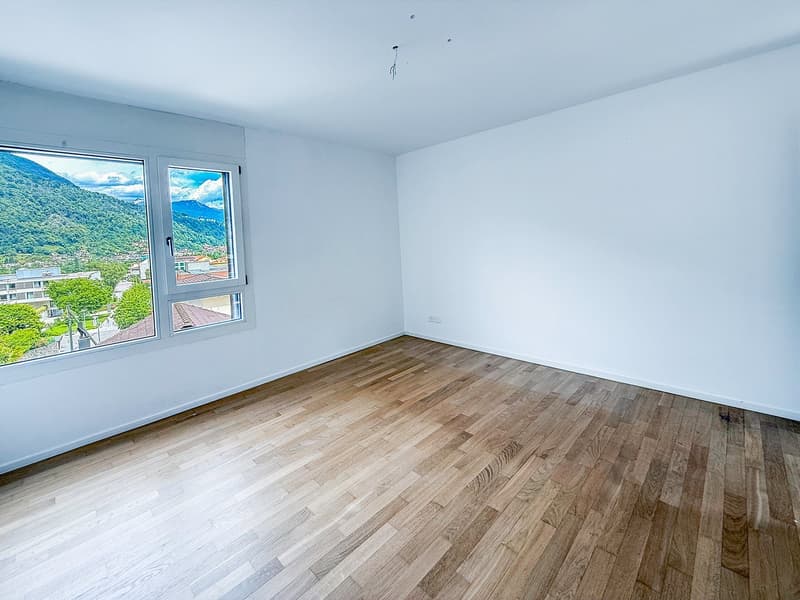 Spacious room with large windows offering a scenic view of the surrounding mountains and landscape. The room has hardwood flooring and white walls, creating a bright and airy atmosphere.