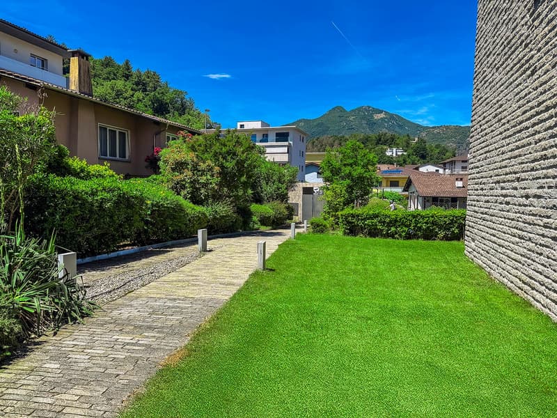 The image shows a residential area with well-maintained landscaping, including a paved walkway, lush green grass, and various shrubs and trees. The buildings in the background appear to be multi-story apartments or condominiums, and there is a balcony or