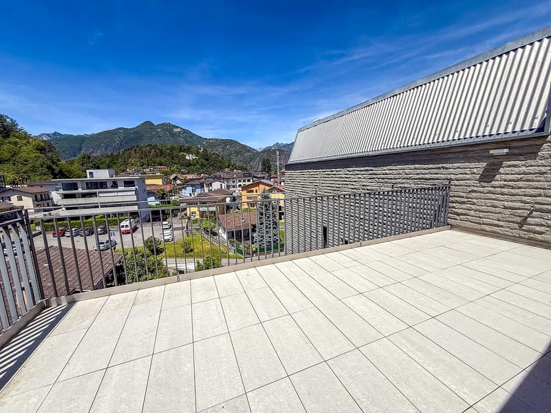 The image shows a balcony or terrace with a view of a town surrounded by mountains. The balcony has a tiled floor and a metal railing. The building has a corrugated metal roof and appears to be part of a larger residential or commercial complex.