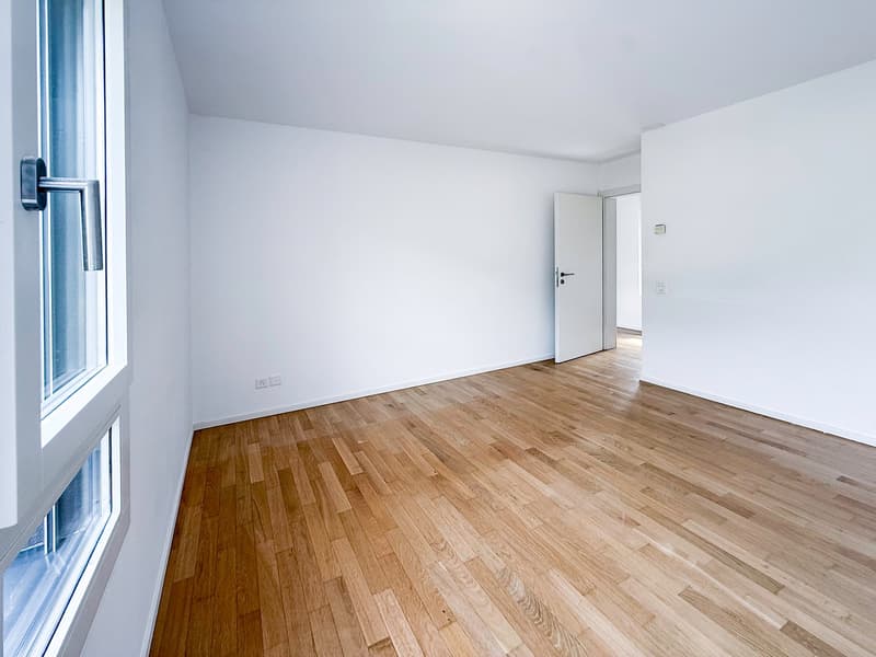 Spacious bedroom with white walls, hardwood flooring, and a large window providing natural light. The room appears to be empty, ready for furnishing and personalization.