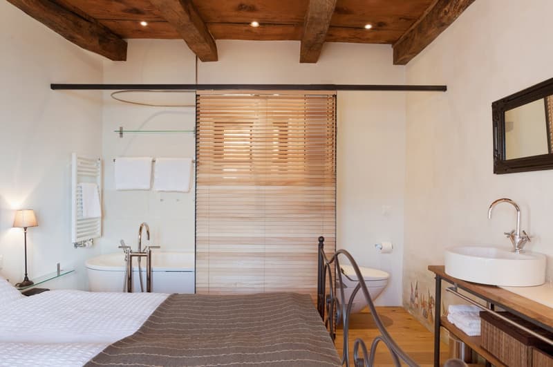 The image shows a bedroom with an en-suite bathroom. The bedroom features a wooden ceiling with exposed beams, a bed with a gray striped blanket, and a wooden vanity with a vessel sink. The bathroom area has a sliding wooden privacy screen, a toilet, and