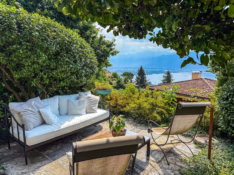 Cozy outdoor patio with a comfortable sofa, chairs, and a scenic view of the lake and mountains in the background, framed by lush greenery and foliage.