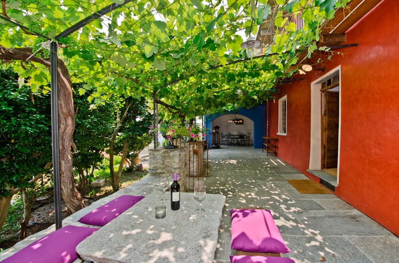 Covered outdoor patio with stone floor, wooden beams, and lush greenery overhead. There is a table with wine glasses and a bottle of wine, as well as comfortable purple cushions on the seating area. The patio leads to a larger covered space with additiona