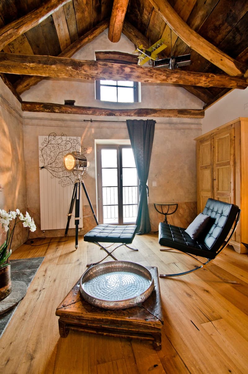 Rustic living room with exposed wooden beams, large windows, leather armchair, tripod lamp, and a decorative metal bowl on a wooden table