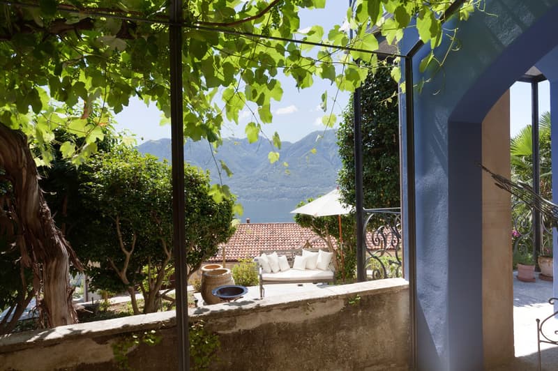 Covered outdoor patio with a seating area overlooking a scenic lake and mountains in the background. The patio is surrounded by lush greenery and trees, creating a peaceful and serene atmosphere.