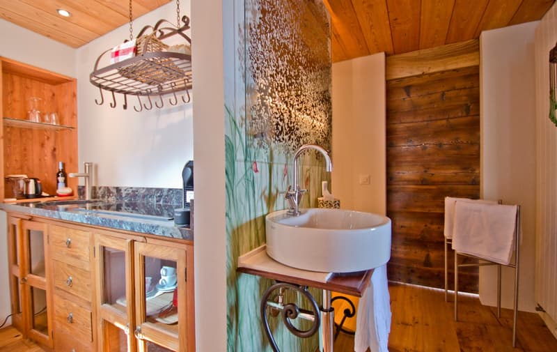 The image shows a rustic kitchen and bathroom area. The kitchen features wooden cabinets, a marble countertop, and a hanging pot rack. The bathroom area has a vessel sink on a wooden vanity, with a decorative tile wall behind it. The overall style is a mi