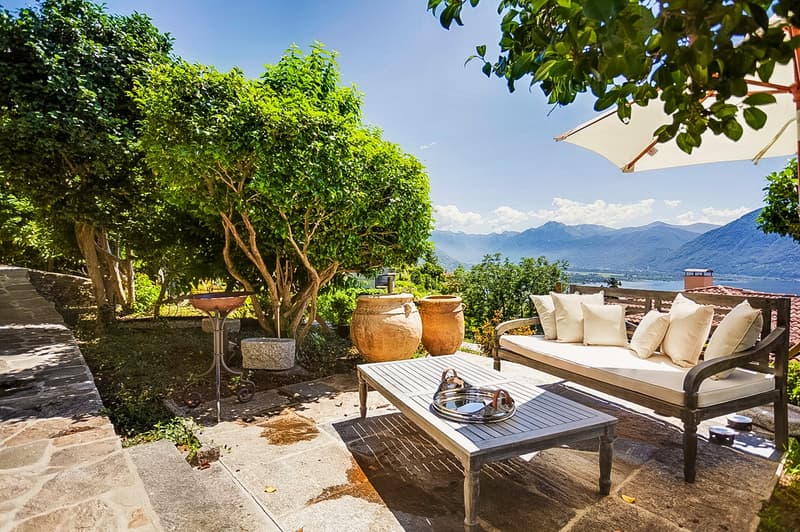 Lush green trees and foliage, stone patio with wooden furniture, large outdoor umbrella, scenic mountain view in the background