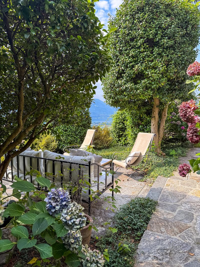 Lush, green garden with trees, shrubs, and flowering plants. Two lounge chairs on a stone patio overlooking a scenic view of the lake or mountains in the distance.