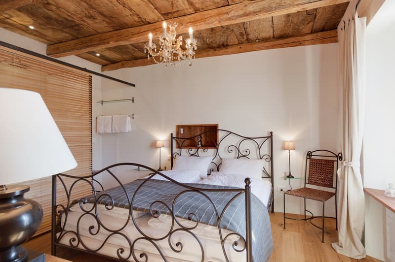 Rustic bedroom with exposed wooden ceiling beams, a wrought-iron bed frame, a chandelier, and a fireplace. The room has a cozy and elegant feel with white bedding and curtains, and a wicker chair in the corner.