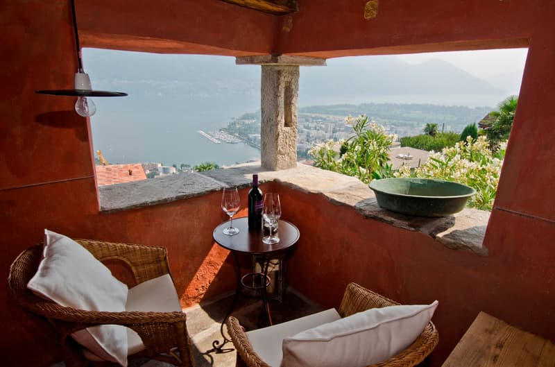 Cozy outdoor seating area with wicker chairs, a small table with wine glasses and a bottle of wine, overlooking a scenic view of the city and water below. The area is surrounded by stone walls and has potted plants and flowers adding to the ambiance.