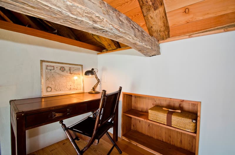 The image shows a cozy home office space with exposed wooden beams on the ceiling, a wooden desk with a lamp and framed artwork, and a wooden bookshelf with decorative items. The room has a warm, rustic feel with the natural wood elements and the presence