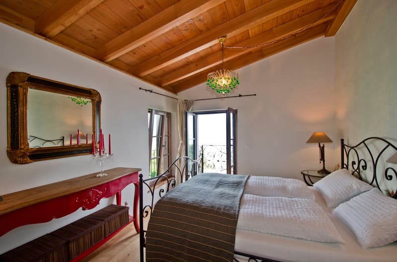 Spacious bedroom with wooden ceiling, large windows, ornate mirror, red dresser, iron bed frame, and a chandelier with green accents.