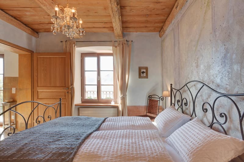 Rustic bedroom with wooden ceiling beams, a crystal chandelier, a balcony with wrought iron railings, and a fireplace. The room features a metal bed frame with a patterned bedspread and pillows, a wooden dresser, and a window overlooking the outdoor scene