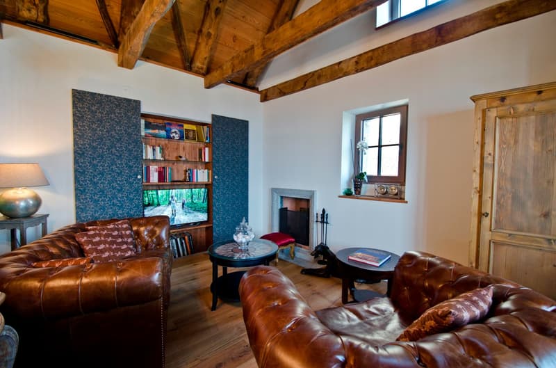 Spacious living room with high wooden beamed ceiling, hardwood floors, leather tufted sofas, a fireplace, a bookshelf, and a window overlooking a forested area