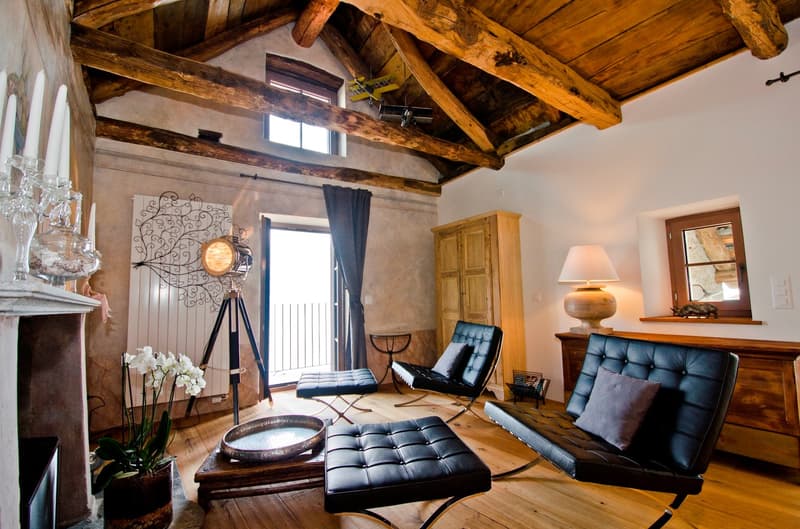 Rustic living room with exposed wooden beams on the ceiling, leather armchairs and ottoman, a wooden cabinet, and a decorative floor lamp. The room has a cozy and warm atmosphere.