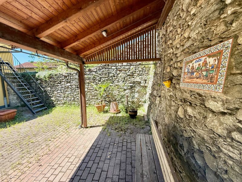 A wooden patio with brick flooring, stone wall, decorative tiles, and hanging lanterns. There are stairs leading to a higher level, and a metal railing on the left side. A small garden with potted plants is present. On the left, a small red pot is placed