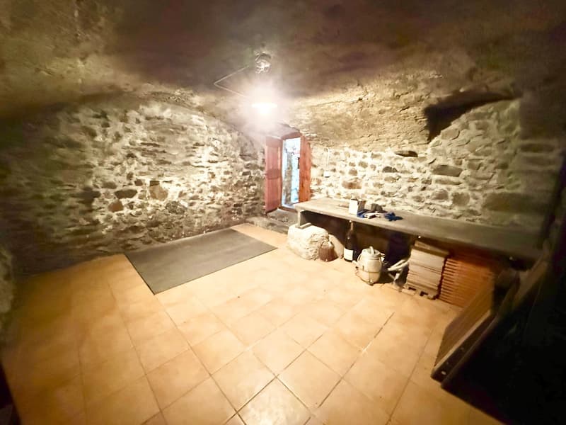 room with stone walls, light on the ceiling, tile floor, wooden bench, desk, bottle and jug, stack of wood, open door leading to a room