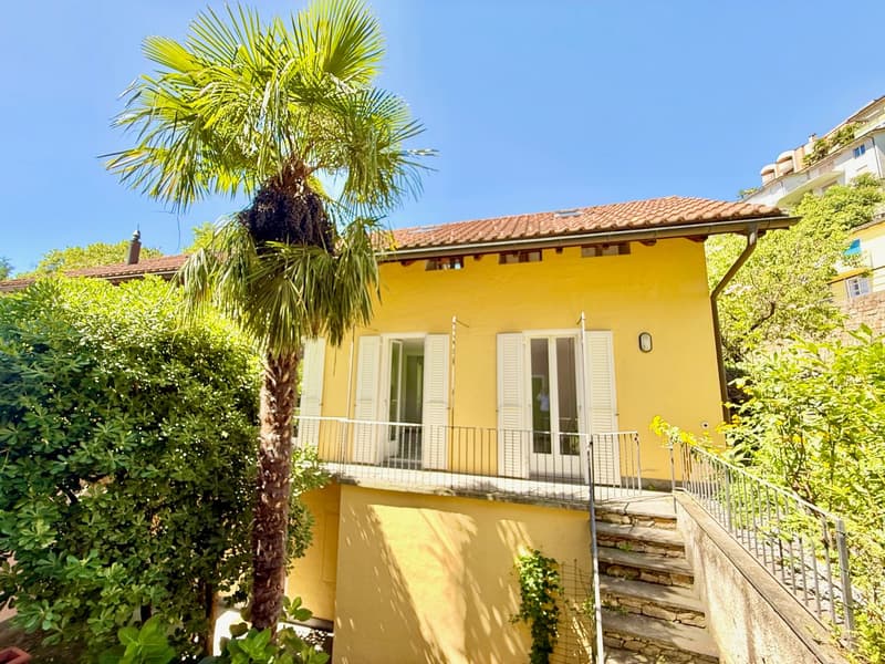 2 story yellow house, red roof, balcony, windows with white shutters, steps leading to entrance, surrounded by trees and plants