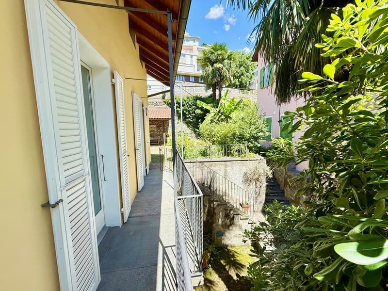 Corridor with tiled floor, two windows, white shutters, a balcony, and a metal railing on the side. The wall is painted yellow. There is a garden with various plants and a building with a balcony and a tree in the distance.