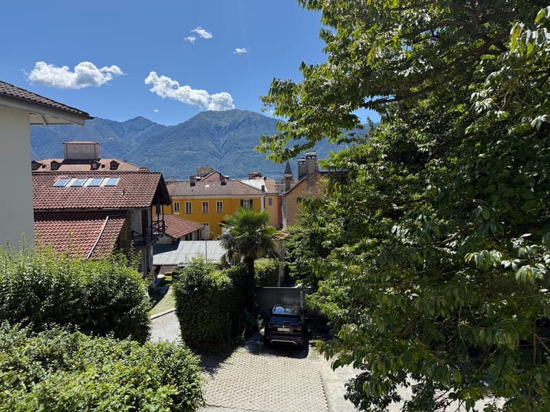 Houses with red roofs, yellow house, mountains in the background, car parked in a garage, driveway, trees and bushes, clear sky with clouds