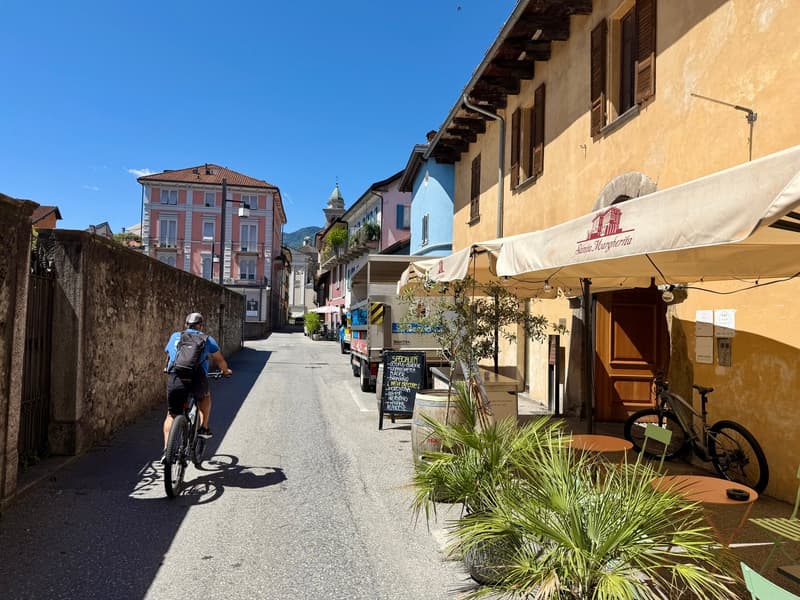 Narrow street with multistory buildings on both sides, a cyclist with a backpack, outdoor tables and chairs under awnings, potted plants, bicycles parked in front of the building, outdoor menu boards, parked truck on the side of the road.