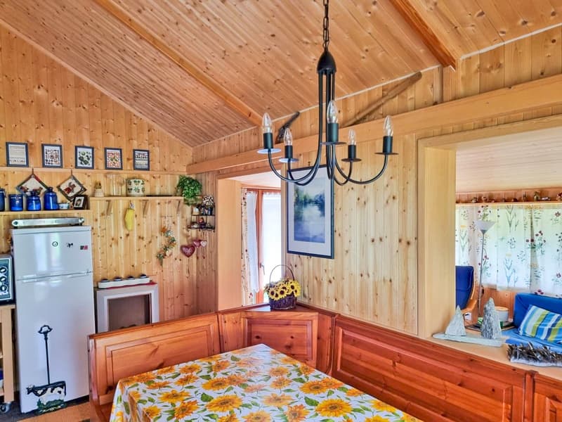 A wooden interior room with a table, refrigerator, microwave, wooden benches, framed pictures on the wall, a window, and a chandelier