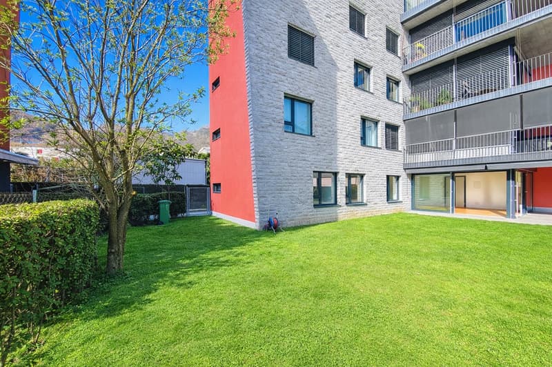 Modern apartment building with red and white facade, balconies, and a well-maintained green lawn in front.