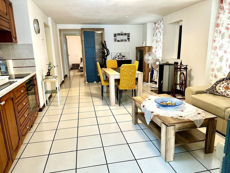 Modern furnished living room and dining room with wood-style flooring, wooden dining table, and blue chairs, a couch in beige color, two fans, wooden kitchen cabinets, stove, microwave, and a table with a potted plant.
