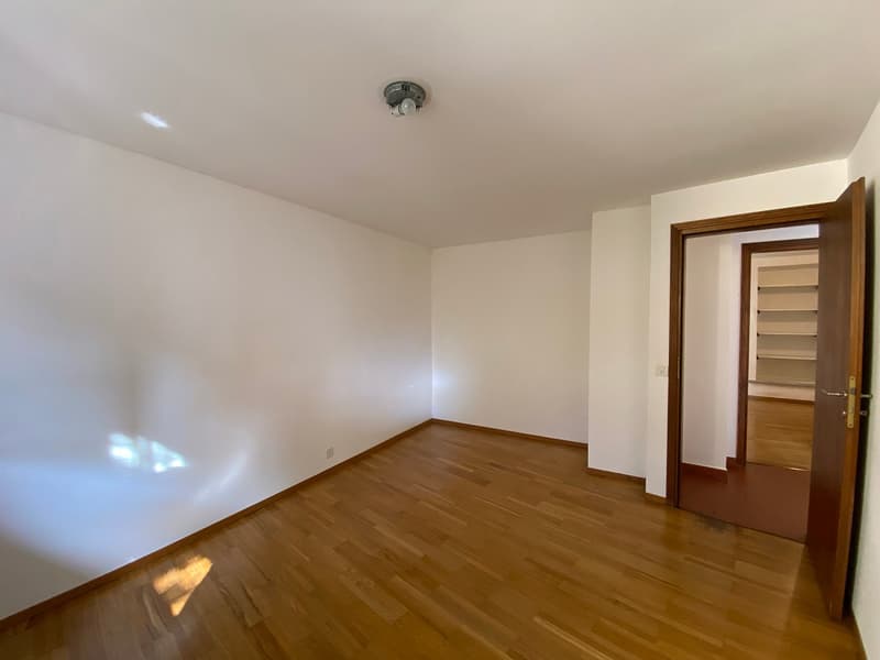Empty room with wooden floor, white walls, ceiling light, open door leading to another room with shelves