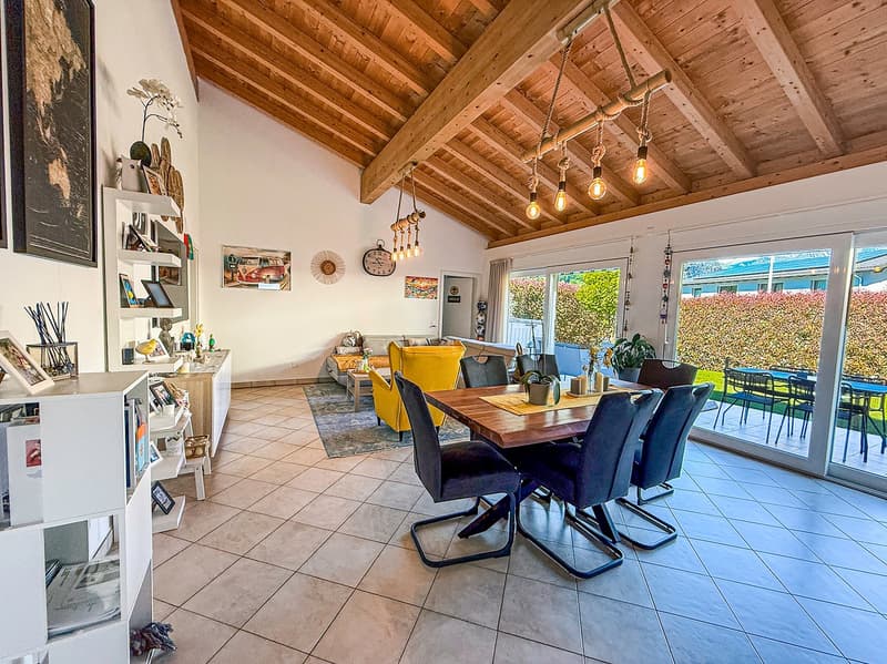 Open concept dining room and living area with wooden ceiling, large table, yellow armchairs, white shelves, dining table with chairs, large glass doors, wooden floor, and view of outside