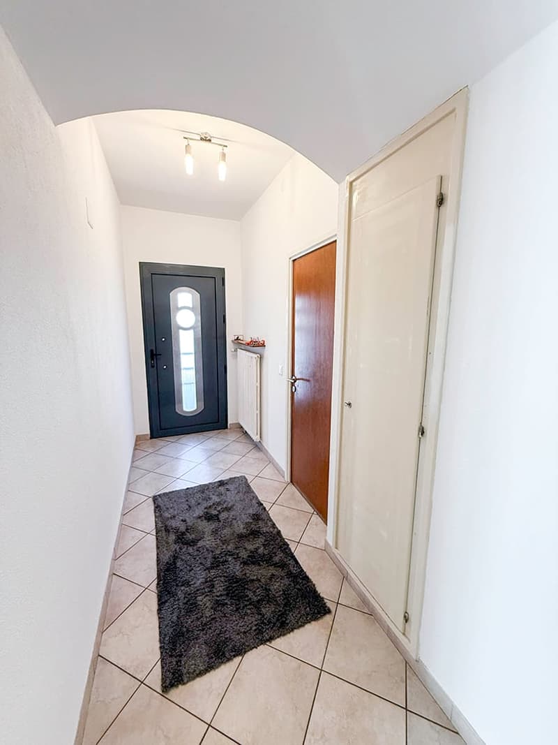 Tiled flooring, white walls, arched ceiling, black front door with window, wooden door to the side, light fixture overhead